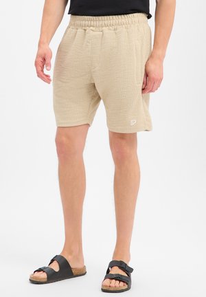 Man draagt beige textuursjorts met elastische taille en zwarte sandalen met gesp, staand tegen een effen witte achtergrond.