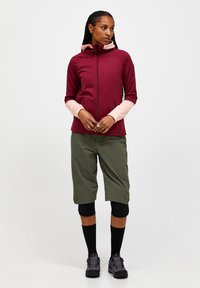 Bordeauxrode en roze ritsjack met capuchon, gecombineerd met olijfgroene shorts en zwarte kniekousen. Geweven stof en sportieve snit.