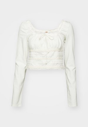 Haut cropped à manches longues blanc avec décolleté carré, détail à cordon, et finition en dentelle sur la poitrine et l'ourlet, présentant une texture froncée.