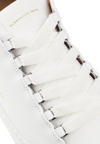 Zapatilla de cuero blanca con cordones planos blancos, ojales plateados y la marca 'Alexander Smith' en relieve. Textura suave con un diseño minimalista.