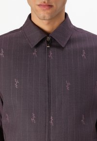 Men's zip-up jacket in dark purple fabric, featuring thin vertical stripes and scattered light pink text logos. Collared design with a smooth finish.