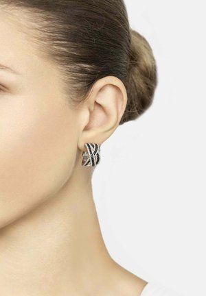 Woman with light skin and hair in a bun wearing a silver and black patterned hoop earring on her left ear against a plain white background.