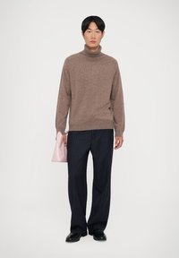 Brązowy sweter z golfem, miękka faktura, w połączeniu z granatowymi spodniami w prążki. Trzyma jasnoróżową torbę, czarne buty dopełniają outfit.