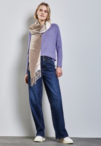 Lila gestrickter Pullover, beige-weißer Schal mit Fransen im Verlauf, dunkelblaue weit geschnittene Jeans und weiße Sneakers, alles vor einem neutralen Hintergrund.