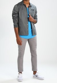 Szara lekka kurtka na niebieskiej koszuli, do tego jasnoszare spodnie o slimowanym kroju i białe trampki z czarnymi akcentami i kropkami.