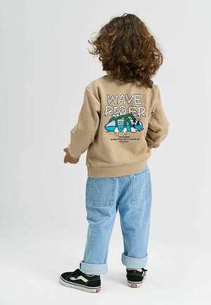 Dítě s kudrnatými vlasy, které má na sobě béžovou mikinu s grafikou "Wave Rider" a světle modré nohavice s ohrnutím, stojící na jednobarevném pozadí.