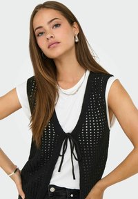 Gilet nero lavorato a maglia con trama aperta, con chiusura a fiocco. Indossato sopra una maglietta bianca, abbinato a jeans scuri e dettagli in gioielli argentati.