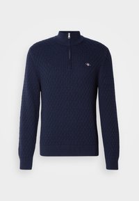 Pull en maille bleu marine avec un motif en losange, manches longues, col à fermeture éclair partielle, et petit logo brodé sur la poitrine.