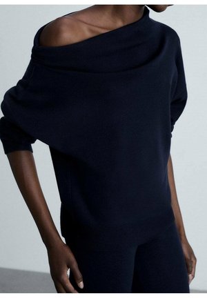 Frau trägt schwarzen Off-Shoulder Langarmpullover und schwarze Hose, steht mit einer Hand nahe der Hüfte vor einem einfarbigen Hintergrund.