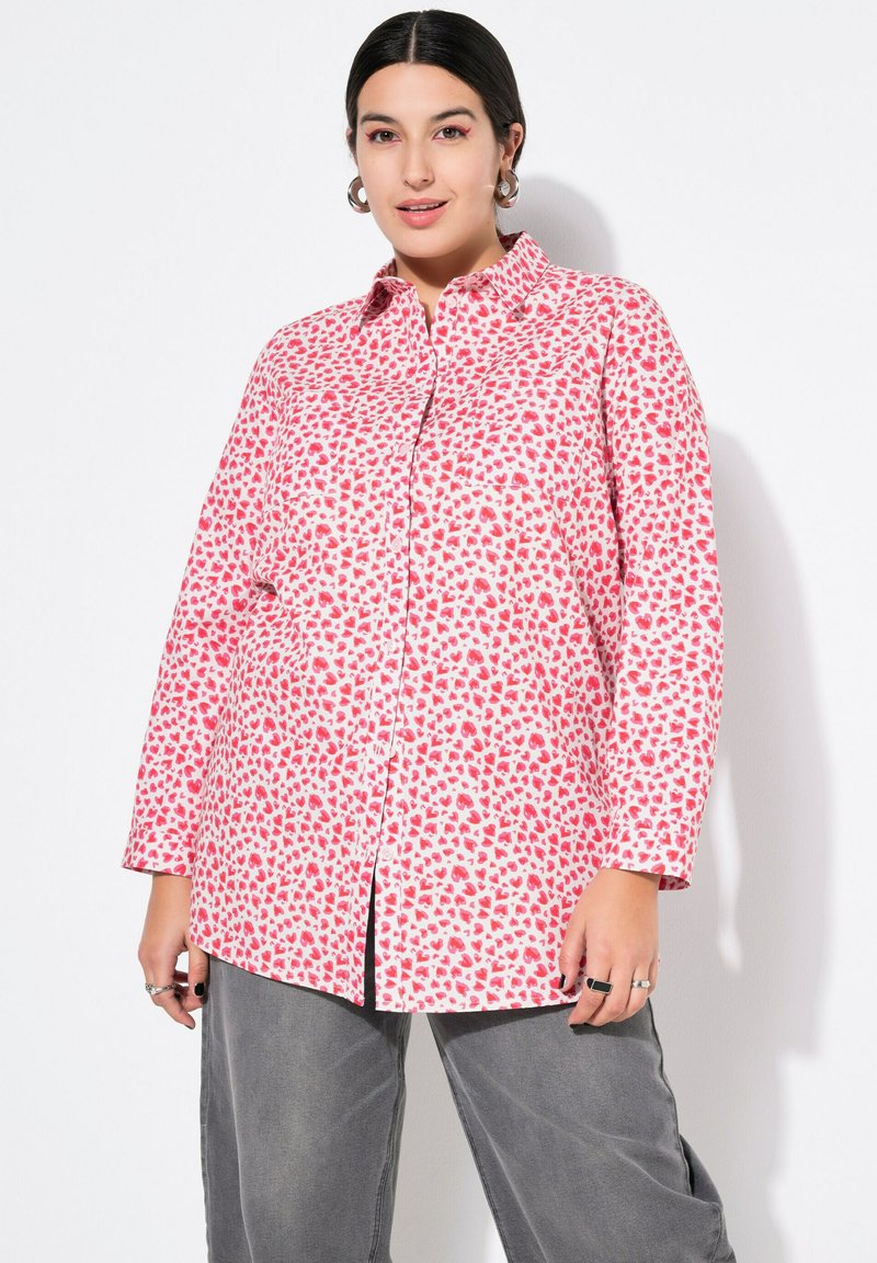Langærmet skjorte i pink stof med hjerte mønster, der har en knap ned design og en afslappet pasform. Grå bukser med en løs skæring.