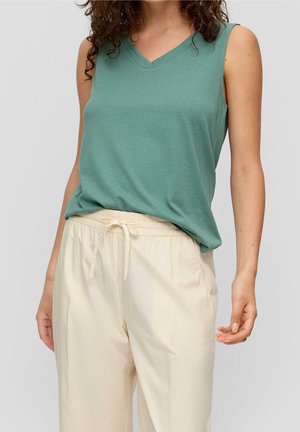 Vrouw draagt een mouwloze groene V-hals top en crèmekleurige broek met trekkoord tegen een effen achtergrond.