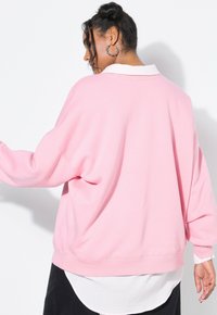 Donna con capelli ricci che indossa orecchini a cerchio, una felpa rosa larga sopra una maglietta bianca e pantaloni neri, rivolta di lato su uno sfondo bianco.