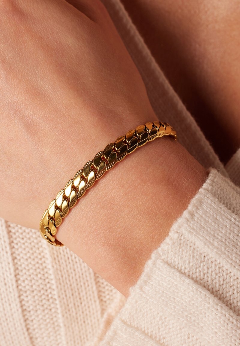 Gouden kettingarmband om een pols, met in elkaar grijpende schakels met een gestructureerd oppervlak, tegen een licht gebreid trui.