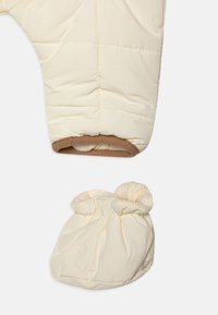 Pilote de neige crème matelassé avec bordure marron et chausson assorti en tissu doux, présentant des bords élastiqués et une texture gonflée.