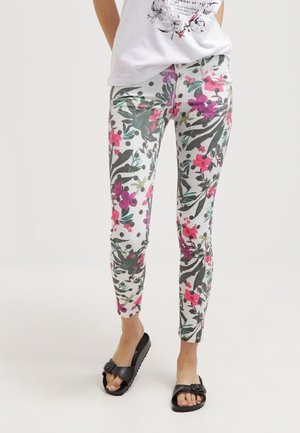 Pantalones blancos ajustados con patrones florales y de hojas coloridos, combinados con una camiseta blanca estampada y sandalias de tiras negras con hebilla.