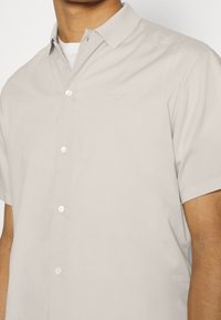 Camicia corta beige chiaro a maniche corte con colletto button-down, realizzata in tessuto liscio, con un ricamo logo discreto sul petto.