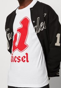 Svart varsityjacka med krämfärgade detaljer, lager på en vit t-shirt med fet röd text och en grafisk logotyp.