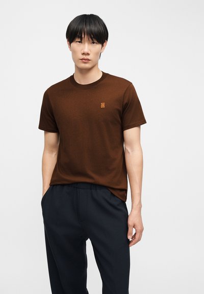 Homme debout, les mains dans les poches, portant une chemise marron à manches courtes avec un petit logo orange et un pantalon bleu foncé, sur fond uni.