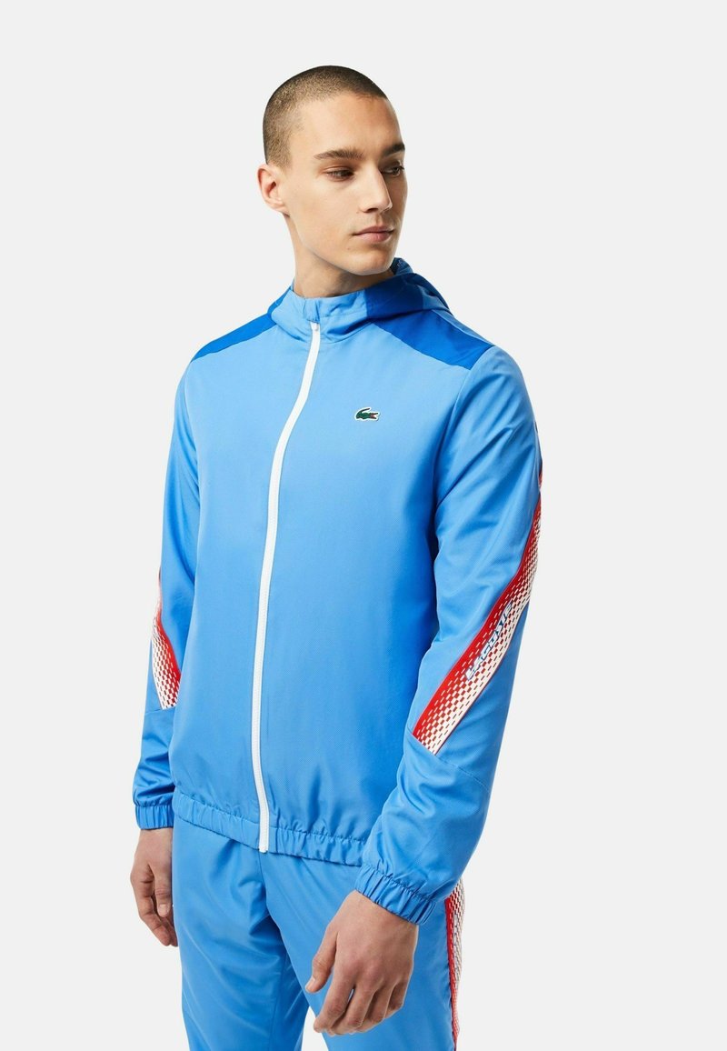 Jeune homme portant un survêtement bleu avec des rayures rouges et blanches sur les manches et le pantalon, se tenant contre un fond blanc.