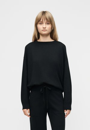 Młoda kobieta o prostych, jasnobrązowych włosach, ubrana w luźną czarną bluzkę z długim rękawem i czarne spodnie ze sznurkiem, stojąca przodem na jednolitym tle.