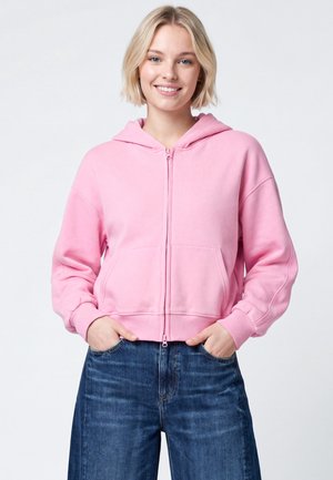Giovane donna sorridente che indossa una felpa rosa con zip e jeans blu con le mani nelle tasche su uno sfondo chiaro e uniforme.