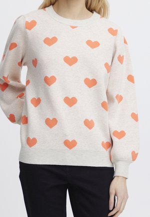 Beżowy sweter z długimi rękawami ozdobiony równomiernie rozmieszczonymi pomarańczowymi wzorami w kształcie serc, noszony przez osobę o jasnych włosach.