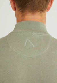 Polo en tissu tricoté vert clair. Présente un col côtelé et un petit détail brodé blanc à l'arrière du col.