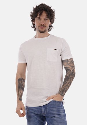 Uomo con capelli ricci che indossa una maglietta bianca con tasche e jeans blu, mostrando tatuaggi dettagliati su entrambe le braccia su uno sfondo semplice.