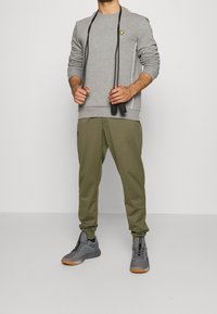 Gray sweatshirt with a small yellow logo, paired with olive green jogger pants. Gray athletic shoes with a gum sole. Black jump rope in hands.