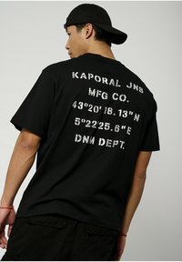 T-shirt en coton noir à manches courtes, avec un grand texte imprimé en blanc au dos, incluant des coordonnées et des détails de la marque.