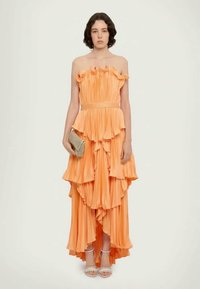 Vestido laranja sem alças com camadas de babados, textura plissada e cintura apertada. Combinado com uma carteira prateada e saltos brancos com tiras.