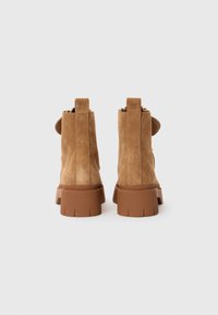 Botines de ante marrón con suela de goma gruesa y detalle de pestaña en la parte posterior, que cuentan con un acabado texturizado y líneas limpias.