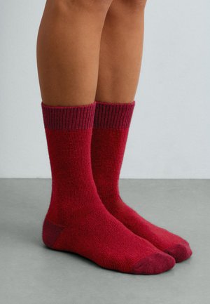 Une personne porte des chaussettes en laine rouges à revers côtelés bordeaux foncé et se tient sur un sol gris, devant un mur clair.