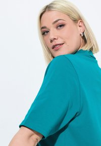 Tyrkysový top s krátkým rukávem vyrobený z hladké tkaniny, s klasickým kulatým výstřihem a přiléhavým střihem, s viditelnými švy.