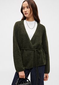 Olivově zelený, texturovaný cardigan s hlubokým výstřihem do V, páskem v pase a dlouhými rukávy, nošený přes bílou blůzu. Drží černou tašku.