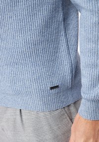 Maglione a coste blu chiaro con superficie testurizzata, dotato di un piccolo etichetta nera sull'orlo, abbinato a pantaloni grigi a quadri.