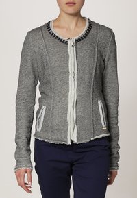 Chaqueta de mezcla de algodón gris con un acabado texturizado, cuello decorativo de cuentas negras, cremallera blanca y dos bolsillos laterales. Detalles de dobladillo deshilachado.