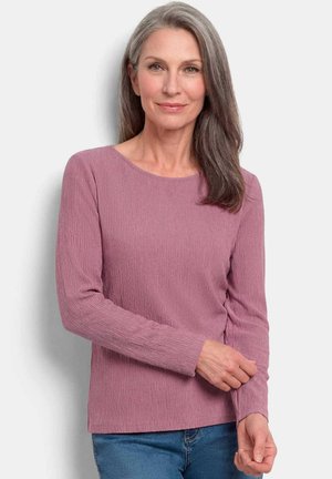Femme d'âge moyen aux longs cheveux gris portant un haut mauve texturé à manches longues et un jean bleu, debout devant un fond clair uni.