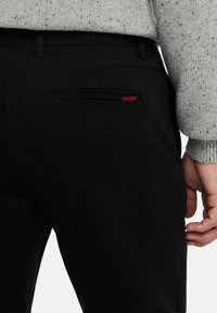 Pantalones negros con una textura suave, que cuentan con un bolsillo trasero con cremallera y un acento rojo. Una mano descansa sobre la costura lateral.