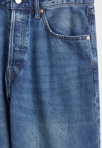 Mėlynos spalvos džinsai su išblukusia tekstūra, penkių kišenių dizainu, variniais vinutėmis ir kontrastingomis siūlėmis palei siūles.