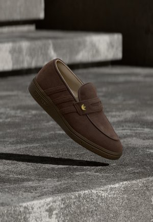 Hnedý semišový mokasín na hladké obúvanie so zlatým logom Adidas, vznášajúci sa nad betónovými schodmi a vrhajúci tieň pod sebou.