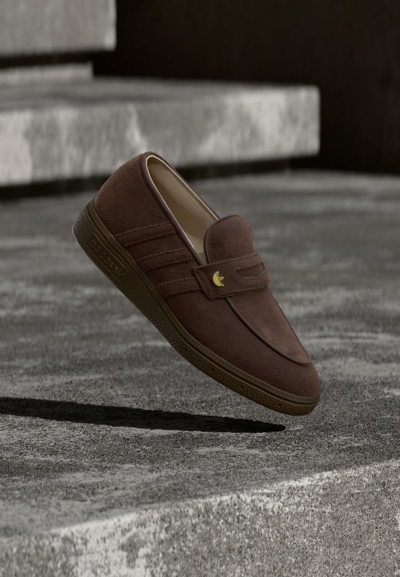 Καφέ σουέντ μοκασίνι slip-on με χρυσό λογότυπο Adidas, αιωρούμενο πάνω από σκάλες από σκυρόδεμα, ρίχνοντας σκιά από κάτω.