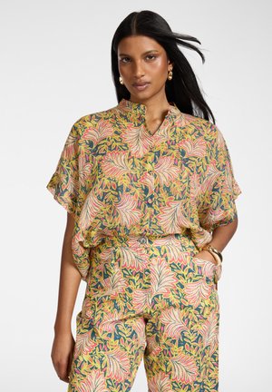 Kobieta nosząca luźną, dopasowaną koszulę i spodnie z żółtym, różowym i zielonym kwiatowym wzorem, złote kolczyki-koła i bransoletki, ręka w kieszeni.