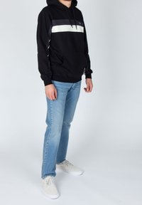 Persona vistiendo una sudadera negra con rayas blancas y grises, jeans azul claro y zapatillas blancas, de pie contra un fondo liso.