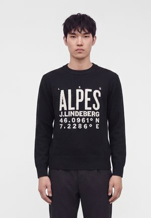 Jeune homme portant un pull noir avec un texte blanc "LES ALPES J. LINDEBERG" et des coordonnées, debout devant un fond clair uni.