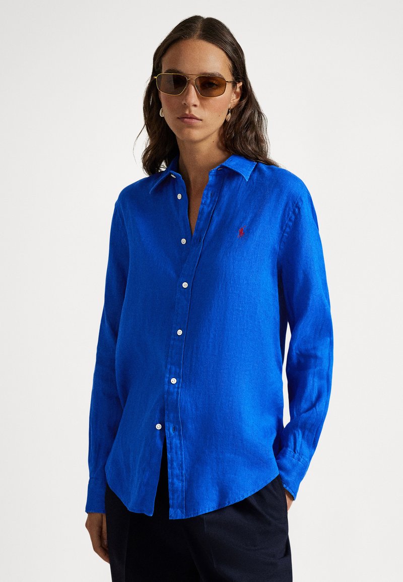 Μπλε λινό πουκάμισο με μακριά μανί κια, κουμπιά μπροστά και μικρό λογότυπο. Υφασμάτινη υφή, άνετη εφαρμογή και στρογγυλεμένη βάση.