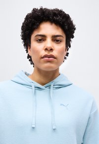 Νεαρό άτομο με κοντά σγουρά μαύρα μαλλιά που φοράει ένα ανοιχτό μπλε υφασμάτινο φούτερ με κουκούλα και το λογότυπο της Puma σε απλό φόντο.