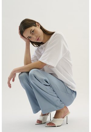 Weißes oversized T-Shirt, hellblaue weit geschnittene Hose und weiße High-Heel-Mules mit pelzigen Akzenten. Subjekt hockt vor einem neutralen Hintergrund.