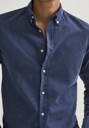 Mężczyzna ubrany w granatową koszulę na guziki, rozpięte dwa górne guziki, ukazujące część szyi i górnej części klatki piersiowej.