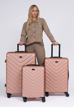 Ensemble de trois valises rigides en rose doré avec un motif en chevron, des accents noirs et des poignées rétractables, positionnées à côté d'une personne.
